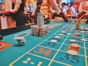 Zahlreiche gestapelte Jetons auf einem Roulette-Tisch eines Casinos; herum viele Spieler und Dealer aktiv.