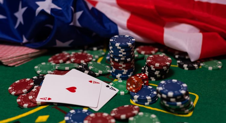 Nahaufnahme von Pokerchips in Rot, Grün und Weiß sowie drei Ass-Karten, die auf einem grünen Filz liegen. Im Hintergrund ist die verschwommene amerikanische Flagge sichtbar.