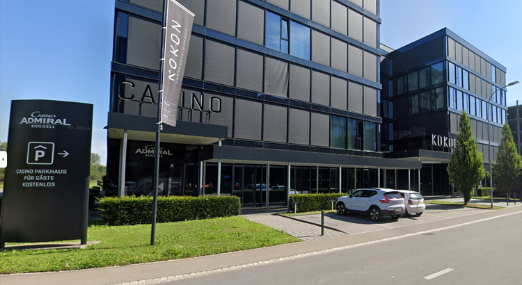 Moderne Außenansicht des Casino Admiral in Ruggell, Liechtenstein, mit grauer Fassade, Glas-Eingang und blau-gelbem Logo, aufgenommen an einem bewölkten Tag. Vor dem Gebäude sind Parkplatzflächen und gepflegte Sträucher sichtbar.