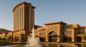 Realistische Darstellung eines modernen Hotel- und Casino-Komplexes mit einer Wasserlandschaft und Springbrunnen im Vordergrund. Die Architektur kombiniert erdige Farbtöne und elegante Linien, während die Fassade des Gebäudes in sanftem Sonnenlicht erstrahlt. Im Hintergrund sind Hügel und ein klarer Himmel zu sehen.