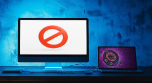 Eine digitale Fotografie zeigt einen Desktop-Monitor mit einem roten „No“-Symbol und ein Laptop, das eine Roulette Szene auf dem Bildschirm zeigt. Die beiden Geräte sind durch ein blaues Ethernet-Kabel miteinander verbunden, während sie auf einem dunklen Holztisch vor einem blauen, texturierten Hintergrund platziert sind