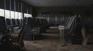 Drei Angestellte stehen vor einer geschlossenen Spielhalle in einer deutschen Stadt. Die Fassade der Spielhalle ist mit heruntergelassenen Rollläden bedeckt, und ein Schild mit der Aufschrift 'SPIELHALLE' ist zu sehen. Die Angestellten tragen Arbeitskleidung und unterhalten sich oder blicken nachdenklich in die Ferne. Die Straße ist leer und das Wetter ist leicht bewölkt