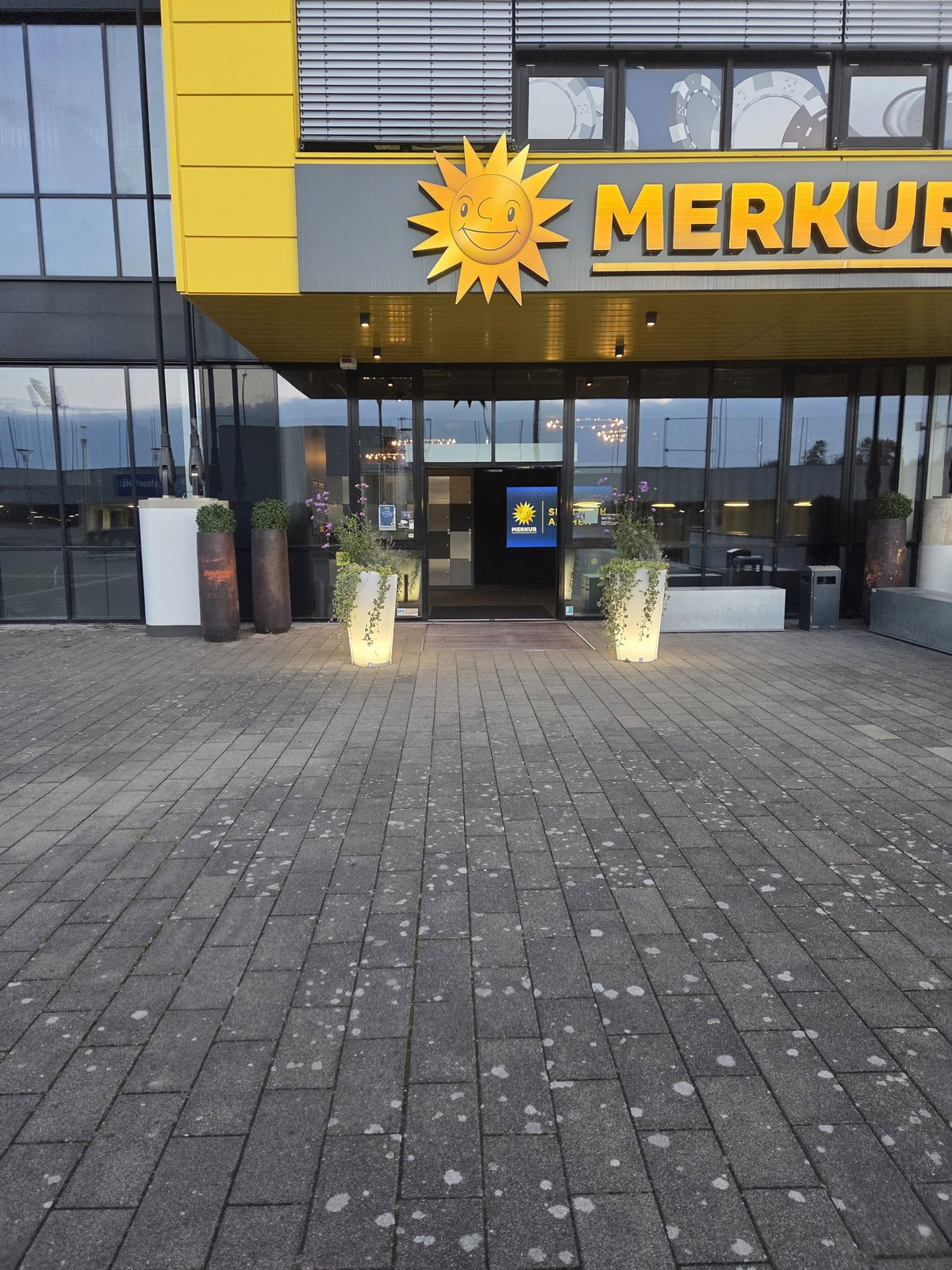 Eingang der Merkur Spielbank Aachen mit Sonnen-Logo und beleuchteten Pflanzkübeln